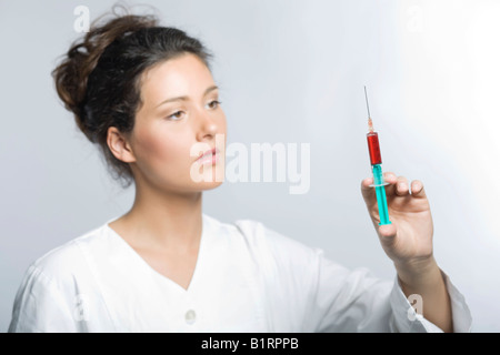 Young woman wearing a white laboratory smock looking concentrated while holding a syringe filled with red liquid in her hand Stock Photo