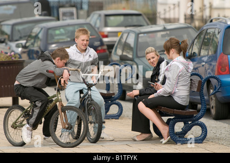 Two teenage boys on bicycles talking to two girls sitting on a bench, UK Stock Photo