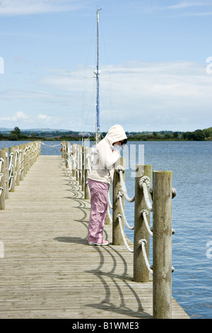 A woman on a pier wearing a hoodie gazes thoughtfully into the water Stock Photo
