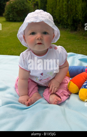 Vertical close up portrait of a baby girl sitting upright in the sunshine in the garden with a sunhat on Stock Photo