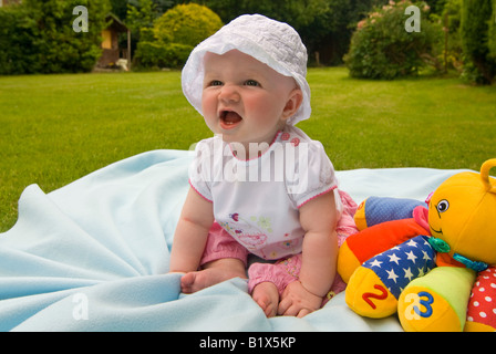 Horizontal close up portrait of a baby girl sitting upright in the sunshine in the garden with a sunhat on Stock Photo