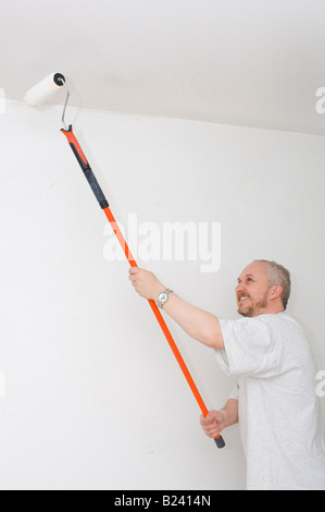 Middle-aged caucasian man with grey hair and a beard painting a ceiling with a paint roller with a long handle, white emulsion paint. Stock Photo