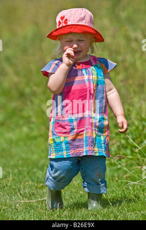 A cute young girl toddler crying wearing wellies and a red sun hat on a sunny day in a green field. Stock Photo