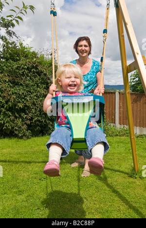 Vertical portrait of an 18 month old little girl having great fun being pushed on her swing by her mother on a sunny day Stock Photo