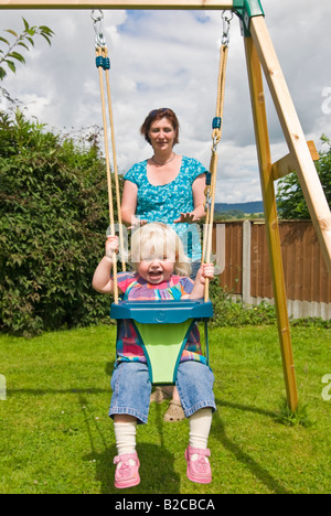 Vertical portrait of an 18 month old little girl having great fun being pushed on her swing by her mother on a sunny day Stock Photo