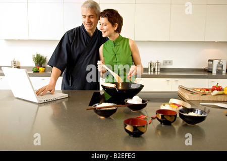 Mid adult woman preparing food with a mature man using a laptop in the kitchen Stock Photo