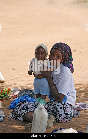 Woman seated on the ground in the shade of a tree waiting for a bus proudly holds up her toddler to pose for the camera Stock Photo
