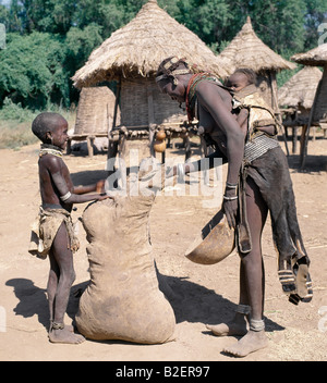 A daughter helps her mother to remove sorghum from a large leather sack. The mother's young baby sleeps  in a leather pouch. Stock Photo