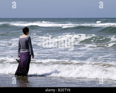 Woman standing in the sea, holding her shoes with the bottom of her skirt in the water. Stock Photo