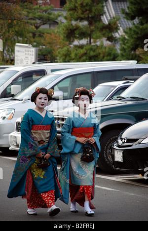 Two Geisha women in colorful, traditional Kimonos stroll past parked cars on their route in downtown Kyoto. Stock Photo