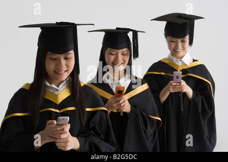 Three young female graduates text messaging with mobile phones and smiling Stock Photo