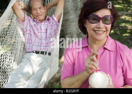 Mature woman holding a coconut milk with her husband lying in a hammock behind her Stock Photo