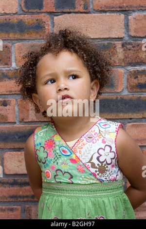 Young mixed race 4yr old girl with curly hair against a brick wall Stock Photo
