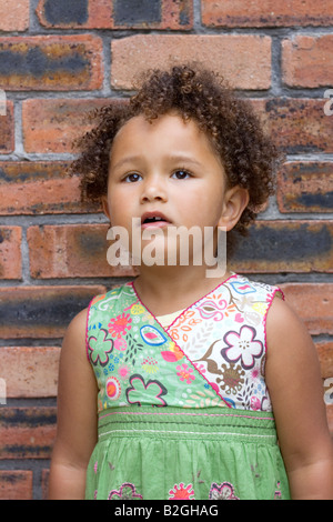Young mixed race 4yr old girl with curly hair against a brick wall Stock Photo