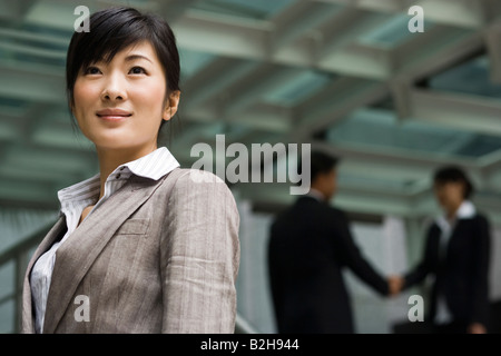 Businesswoman grinning  with two business executives shaking hands behind her Stock Photo