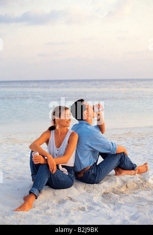 Maldives. Young couple drinking from cans while sitting back to back on sandy beach. Stock Photo