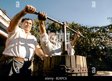 two men pulling pushing turning metal bar conected to mechanism inside a wooden barrel used for pressing grapes Stock Photo