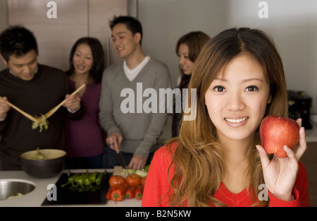 Portrait of a young woman holding an apple while friends working in a kitchen Stock Photo