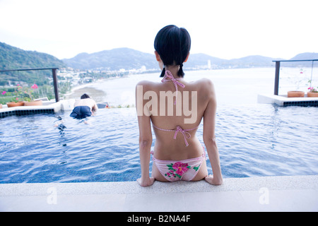 Rear view of a young woman sitting at the poolside with a young man swimming in the background, Phuket, Thailand Stock Photo