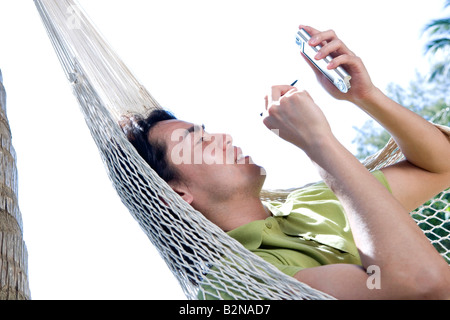 Side profile of a young man reclining in a hammock and using a palmtop, Phuket, Thailand Stock Photo