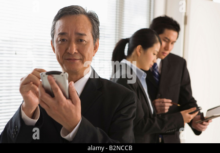 Businessman using a personal data assistant with two business executives standing behind him Stock Photo