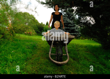 Mother moving her son in a hand wheel barrow through the garden Stock Photo