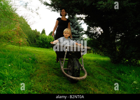 Mother moving her son in a hand wheel barrow through the garden Stock Photo