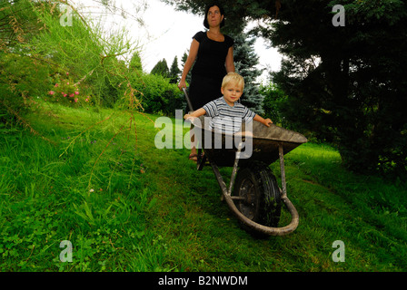 Mother moving her son in a hand wheel barrow through the garden Stock Photo