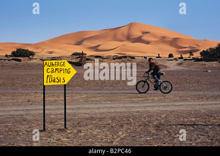 Young boy bicycling past a sign showing the way to a campsite near the Sahara desert near Hassi Labiad in Morocco Stock Photo