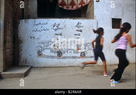 Two girls running past conflict themed graffiti wall in Palestinian Shatila refugee camp,  Beirut, Lebanon, Middle East Stock Photo