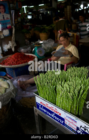 A Bangkok market stall holder sitting by a pile of red chillies and asparagus reading a newspaper Bangkok Thailand Stock Photo