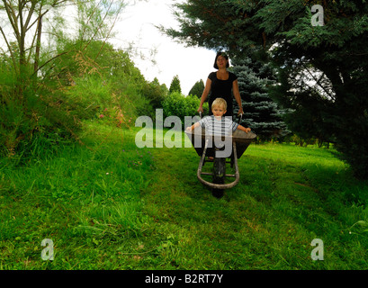 Mother moving her son in a hand wheel barrow through the garden Stock Photo