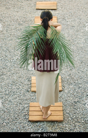 Woman standing on foothpath, carrying palm leaves behind back, rear view Stock Photo