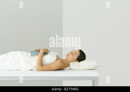 Woman lying on massage table, holding book on chest, eyes closed Stock Photo