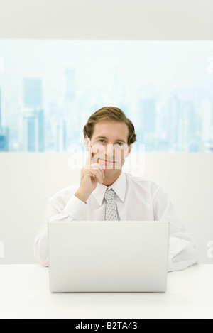 Businessman sitting with laptop computer, holding head, smiling at camera Stock Photo