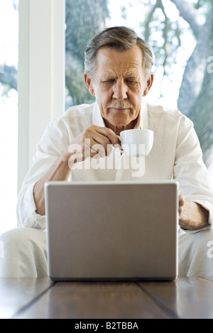 Senior man holding coffee cup, looking down at laptop, sitting with legs apart Stock Photo