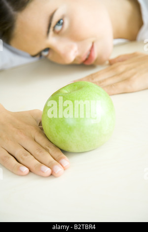 Woman resting head on counter behind apple, looking up, focus on foreground Stock Photo