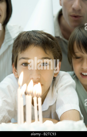 Boy behind birthday cake with lit candles, wearing party hat, surrounded by family, cropped Stock Photo