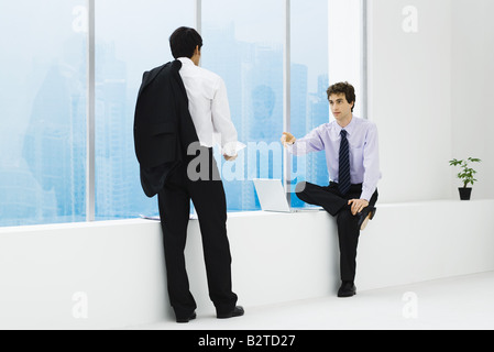 Two businessmen facing each other, one sitting on ledge, one standing Stock Photo
