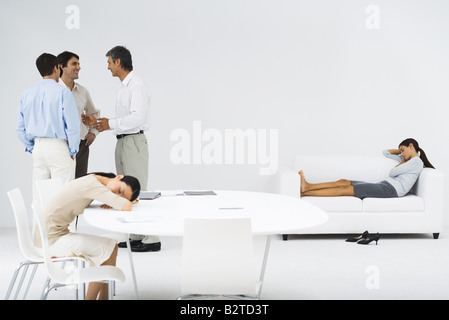 Women napping, one at table, one on couch, men talking in group in the corner Stock Photo