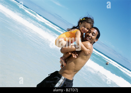 Father carrying daughter at the beach, both smiling at camera