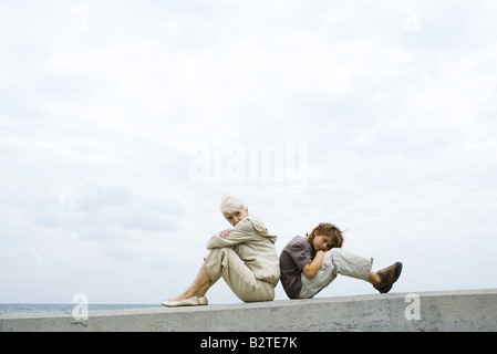 Grandmother and grandson sitting back to back, arms folded, boy looking at camera Stock Photo