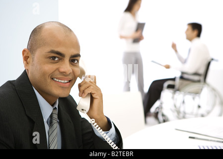 Businessman using phone, smiling at camera, colleagues in background Stock Photo