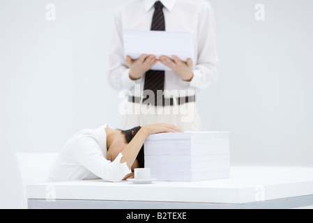 Woman with head down on desk, next to tall stack of papers, man holding another stack of paper Stock Photo