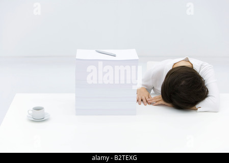 Woman with head down on desk, next to tall stack of papers Stock Photo