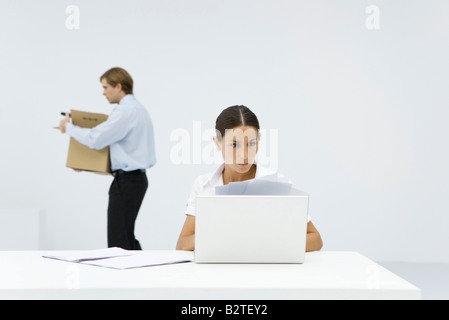 Professional woman reviewing documents at desk behind laptop, colleague carrying box in background Stock Photo