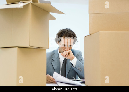 Businessman between stacks of cardboard boxes, studying document, hand over mouth Stock Photo
