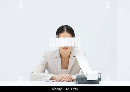 Woman sitting at desk, tape from an adding machine wrapped around her eyes, arms folded Stock Photo