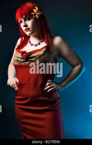 Fashion portrait of a young goth girl with bright red hair in a red dress on a blue background. Stock Photo