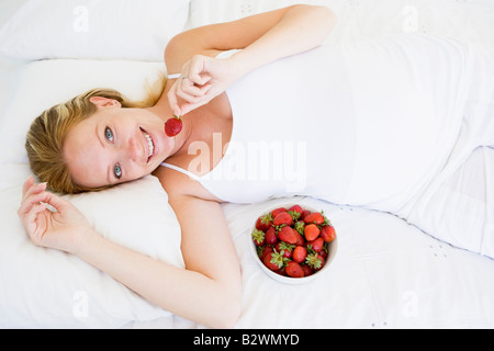 Pregnant woman lying in bed with bowl of strawberries smiling Stock Photo